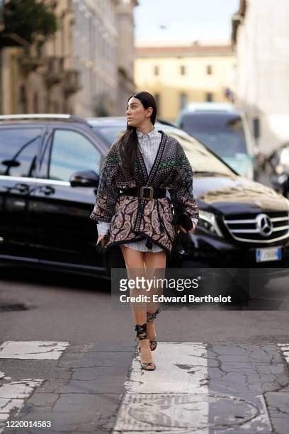Guest wears a long striped shirt, a double breasted long jacket/dress with printed geometric patterns, a large belt, shoes, a black bag, outside...