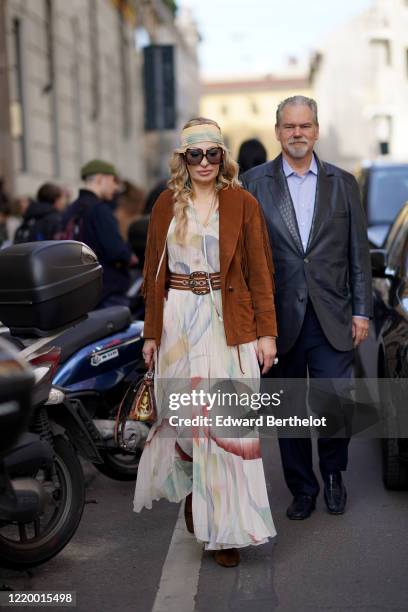 Guest wears a bandanna, a brown suede jacket with fringes, a flowing dress with floral print, a belt, sunglasses, outside Etro, during Milan Fashion...