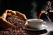 Pouring coffee with smoke on a cup and coffee beans on burlap sack on black background