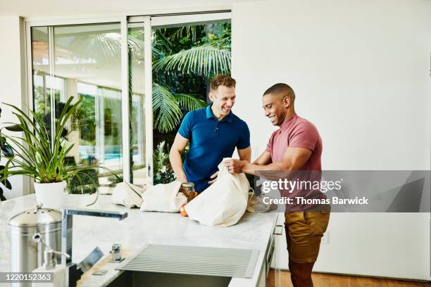 smiling gay couple unloading groceries from canvas bags in kitchen - entladen stock-fotos und bilder