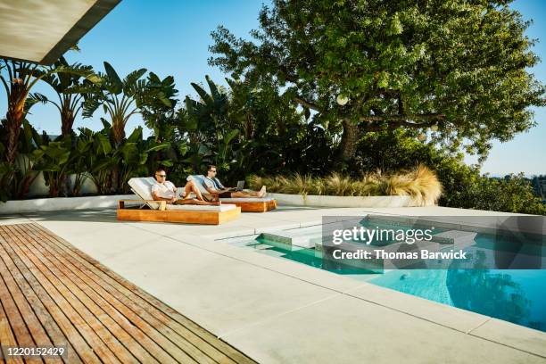 gay couple working on laptop and digital tablet while sitting in lounge chairs by pool - telecommuting couple stock pictures, royalty-free photos & images