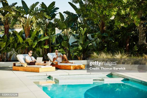 gay couple working on laptop and digital tablet while relaxing by pool - man ipad holiday stockfoto's en -beelden