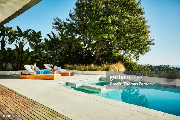 female friends in discussion while relaxing in lounge chairs by pool - friends poolside stock pictures, royalty-free photos & images