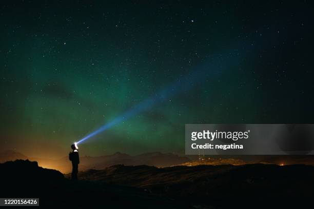 young man with the flashlight on the head stands on the left under the green aurora borealis lighting the ray of light up to the night sky - noorderlicht sterren stockfoto's en -beelden