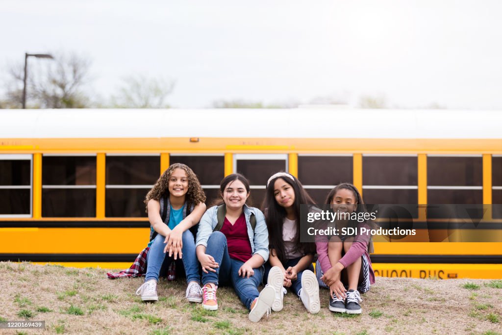 Girls, sitting on grass near school bus, pose for camera