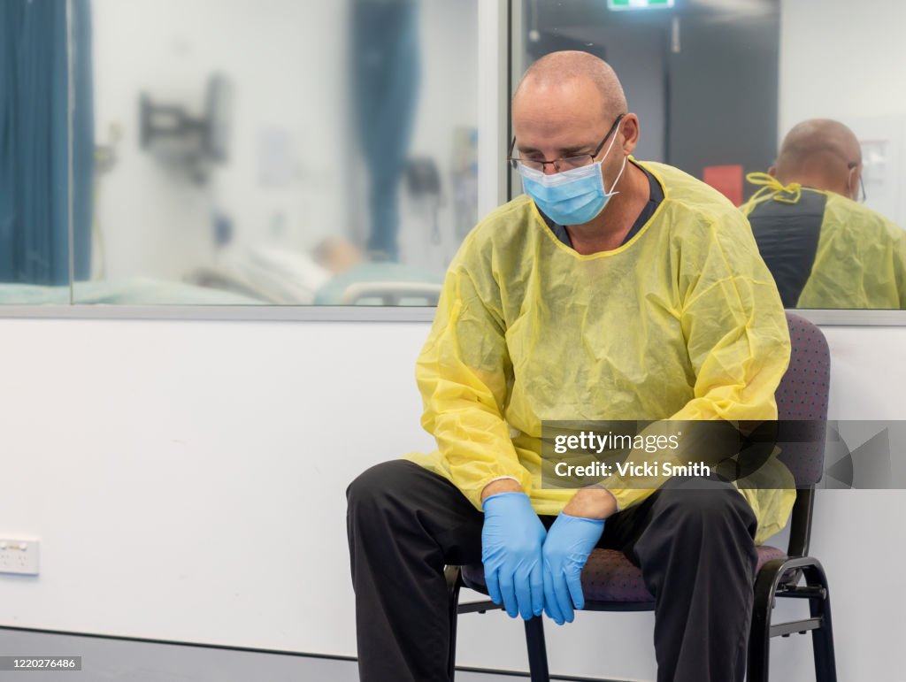 Male Health care sitting in a chair beside a closed hospital room looking exhausted