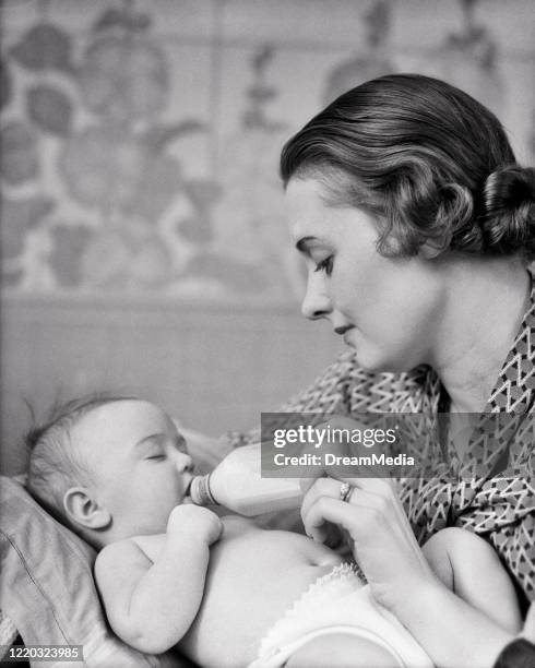 1930s 1940s brunette woman mother feeding holding glass milk bottle for sleepy infant baby girl daughter in nursery