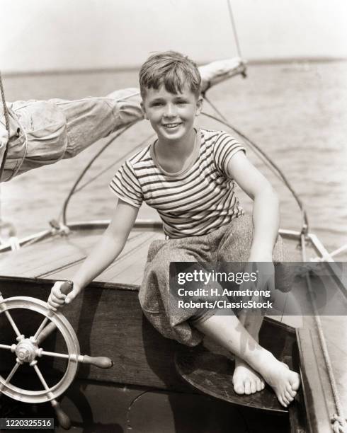 1930s smiling towheaded enthusiastic barefooted boy sitting at the spoked cast iron ships wheel steering piloting small sailboat