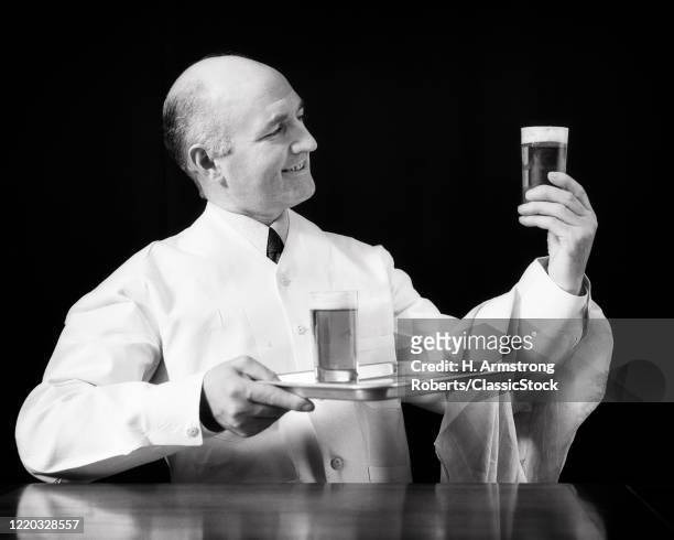 1930s bartender waiter wearing linen jacket uniform holding tray with glass of beer looking at second glass checking the foam