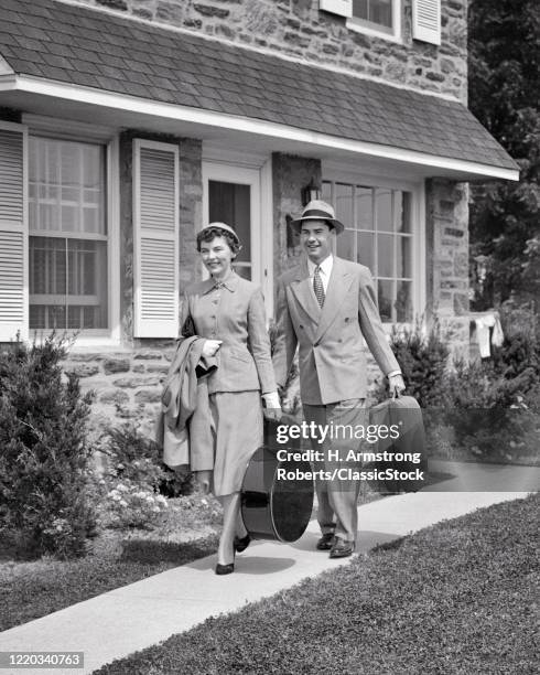 1950s smiling couple walking together carrying luggage leaving suburban house going on vacation trip