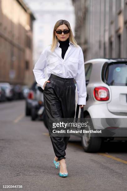 Guest wears sunglasses, a black turtleneck pullover, a white shirt, black leather pants, blue shoes, a Prada bag, outside Sportmax, during Milan...