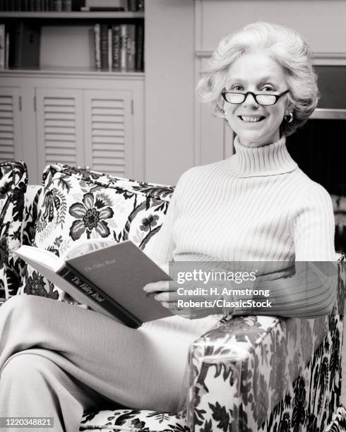 1970s MATURE RELAXED SLENDER HEALTHY WOMAN HOLDING A BOOK WEARING READER EYEGLASSES TURTLENECK SWEATER LOOKING AT CAMERA SMILING