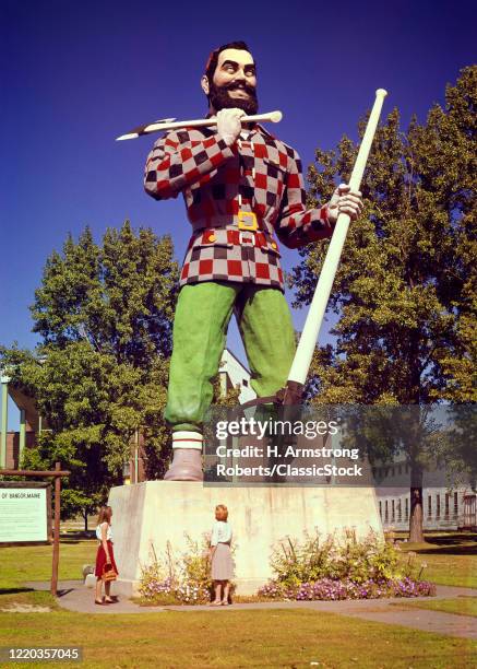 1960s two anonymous tourists back view looking at paul bunyan statue 31 foot tall holding an ax and peavey bangor maine USA