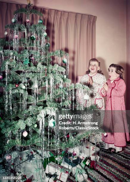1950s two girls standing in front of christmas tree with look of wonder wearing pajamas