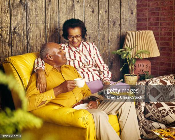 1970s 1980s mature african-american couple sitting on yellow leather chair drinking coffee holding magazine wood paneled room