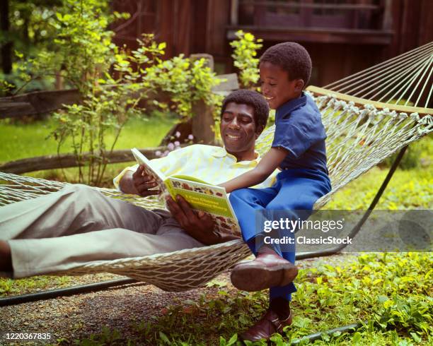 1970s smiling african-american man in a backyard hammock reading a book with his son