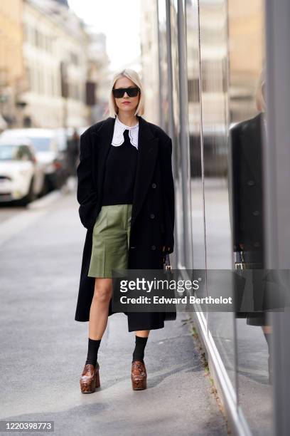 Linda Tol wears sunglasses, an oversized black long jacket, a bag, white large collar, a black top, green shorts, socks, brown leather shoes, outside...