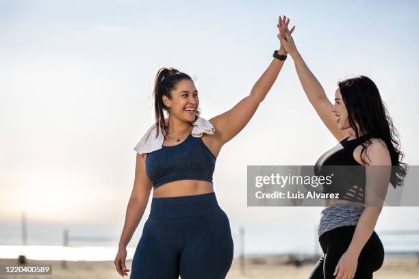 women giving each other a high five after exercising at the beach - formes généreuses photos et images de collection