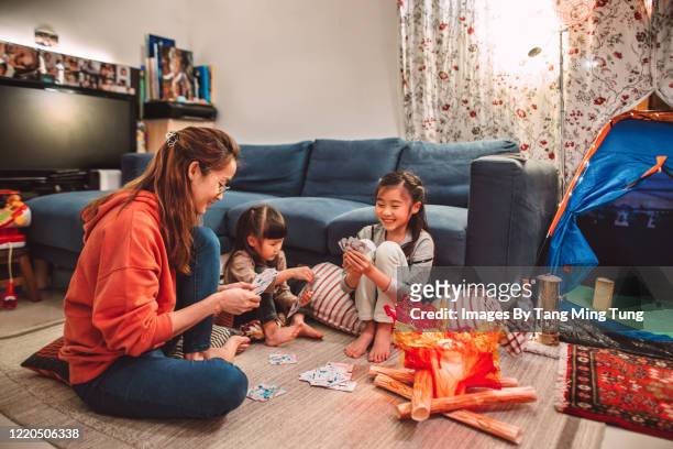 mom & daughters playing card games joyfully at home with a hand crafted fake campfire & camping tent setting besides them in the evening. - camping games stockfoto's en -beelden