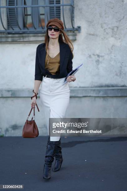 Guest wears a brown beret hat, a navy blue jacket, a brown top, a belt, a white slit skirt, black leather boots, a brown bag, outside Prada, during...