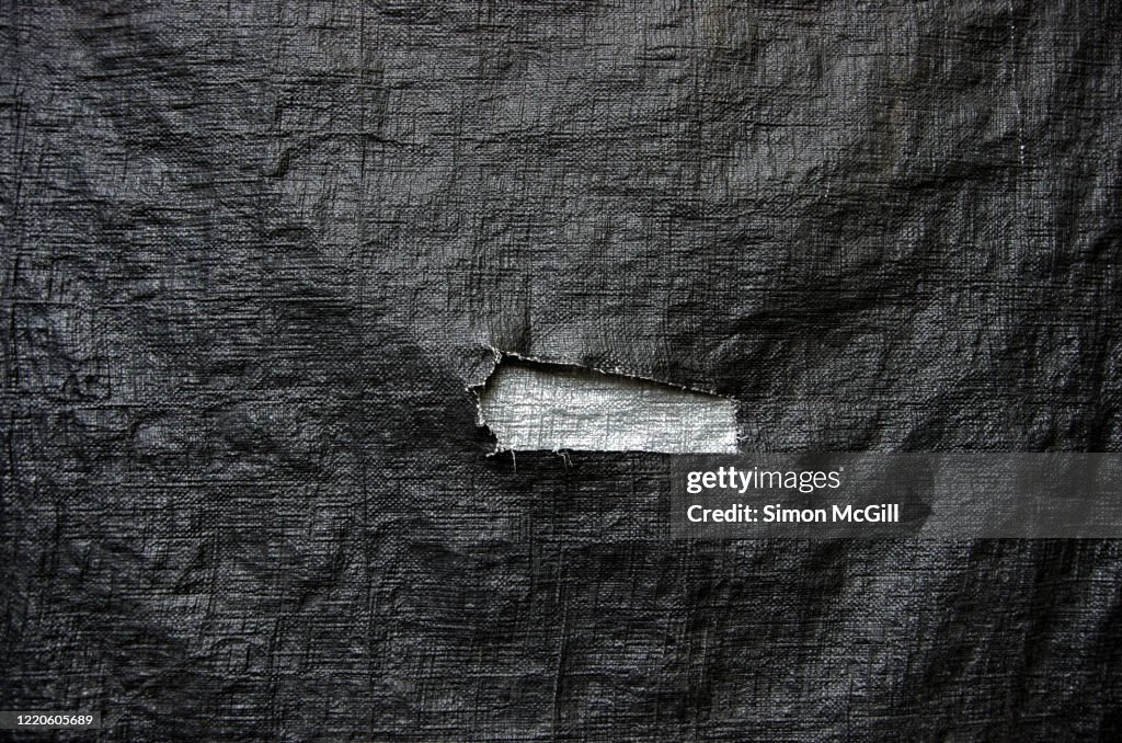 Jagged rectangular tear in a black plastic tarp exposing a silver coloured tarp