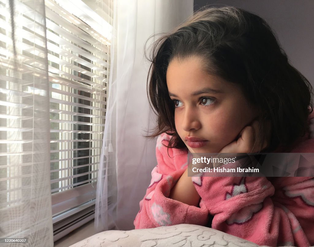 Beautiful 7 years old girl looking through window blinds during the coronavirus epidemic lockdown.