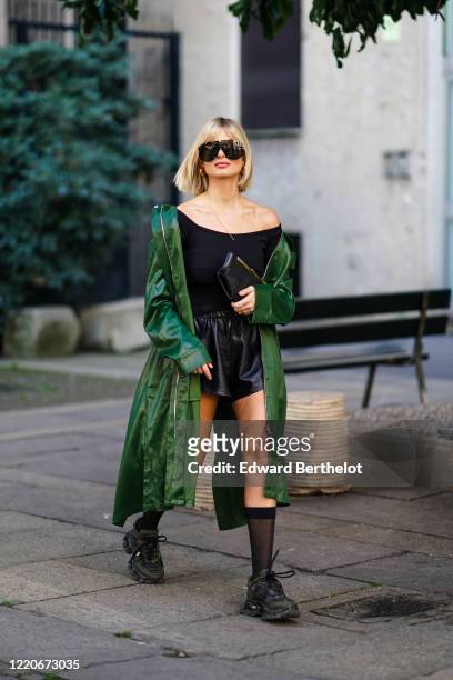 Xenia Adonts wears sunglasses, a black off-shoulder top, a leather short skirt, a green leather coat, a clutch, Prada black socks, black shoes,...