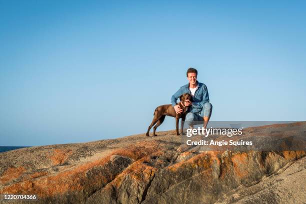 young man crouching and smiling next to labrador dog on rocks at the beach - portrait lachen stock pictures, royalty-free photos & images