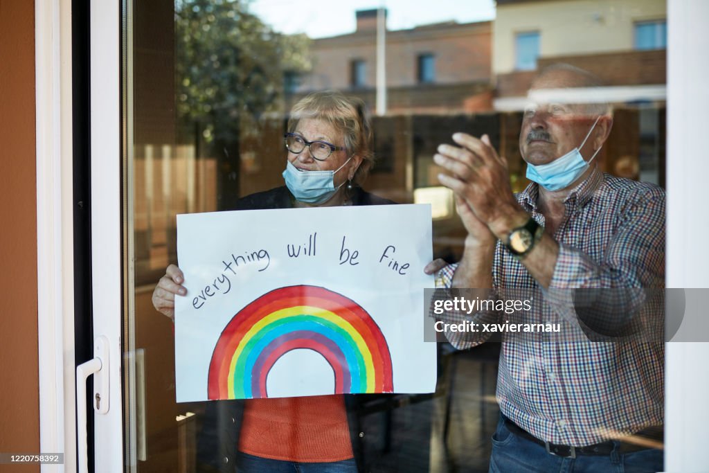 Senior couple on their 70s clapping hands at showing a hand drawn rainbow at home in quarantine