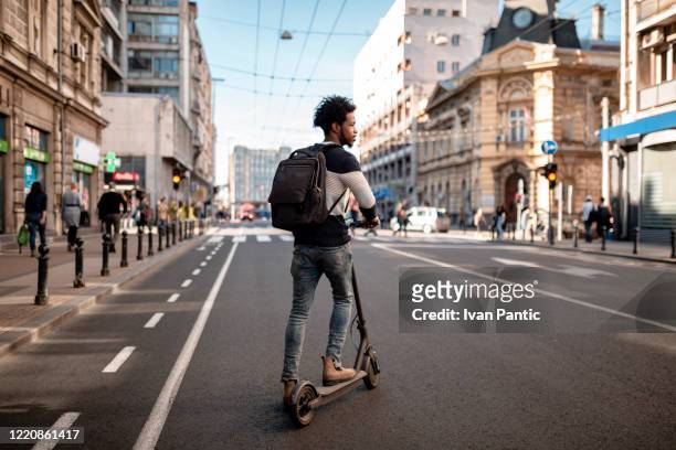 junger mann mit lockigem haar fährt einen elektrischen push-scooter durch die stadt - tretroller stock-fotos und bilder