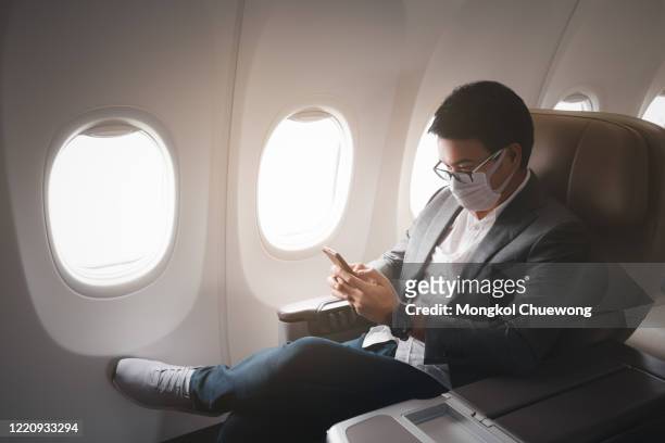 young asian businessman wearing protective face mask with suit sitting in business first class seat using smartphone due to coronavirus or covid-19 outbreak situation in all of landmass in the world - aircraft wifi fotografías e imágenes de stock