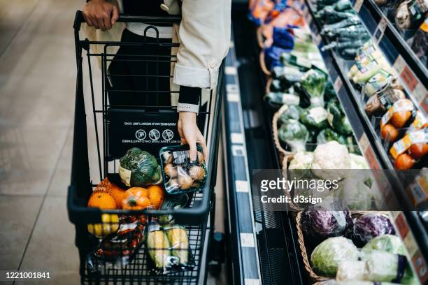 close up of woman's hand shopping for fresh groceries in supermarket and putting a variety of organic vegetables in shopping cart - shopping cart groceries stockfoto's en -beelden