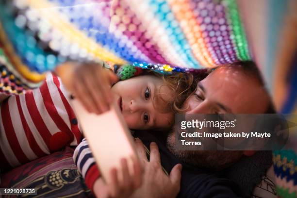boy and dad  watching something in a smartphone under a colorful blanket - at home stock-fotos und bilder