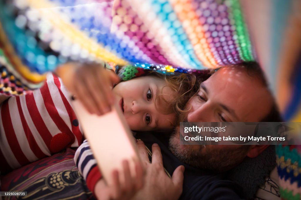 Boy and dad  watching something in a smartphone under a colorful blanket