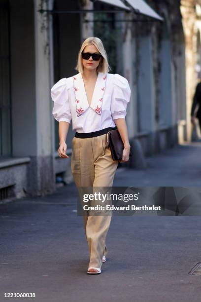 Linda Tol wears sunglasses, a white shirt with ruffled puff shoulders and red floral print, beige flare pants, a brown leather clutch, white shoes,...