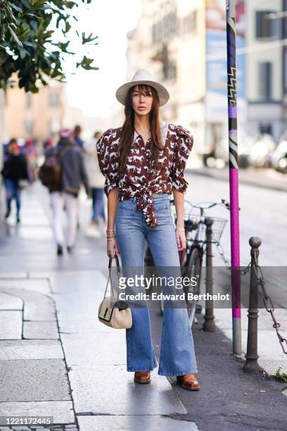 Estelle Chemouny wears a white hat, a ruffled puff shoulders brown shirt with printed horses, blue flare denim jeans pants, a Saint Laurent YSL bag,...