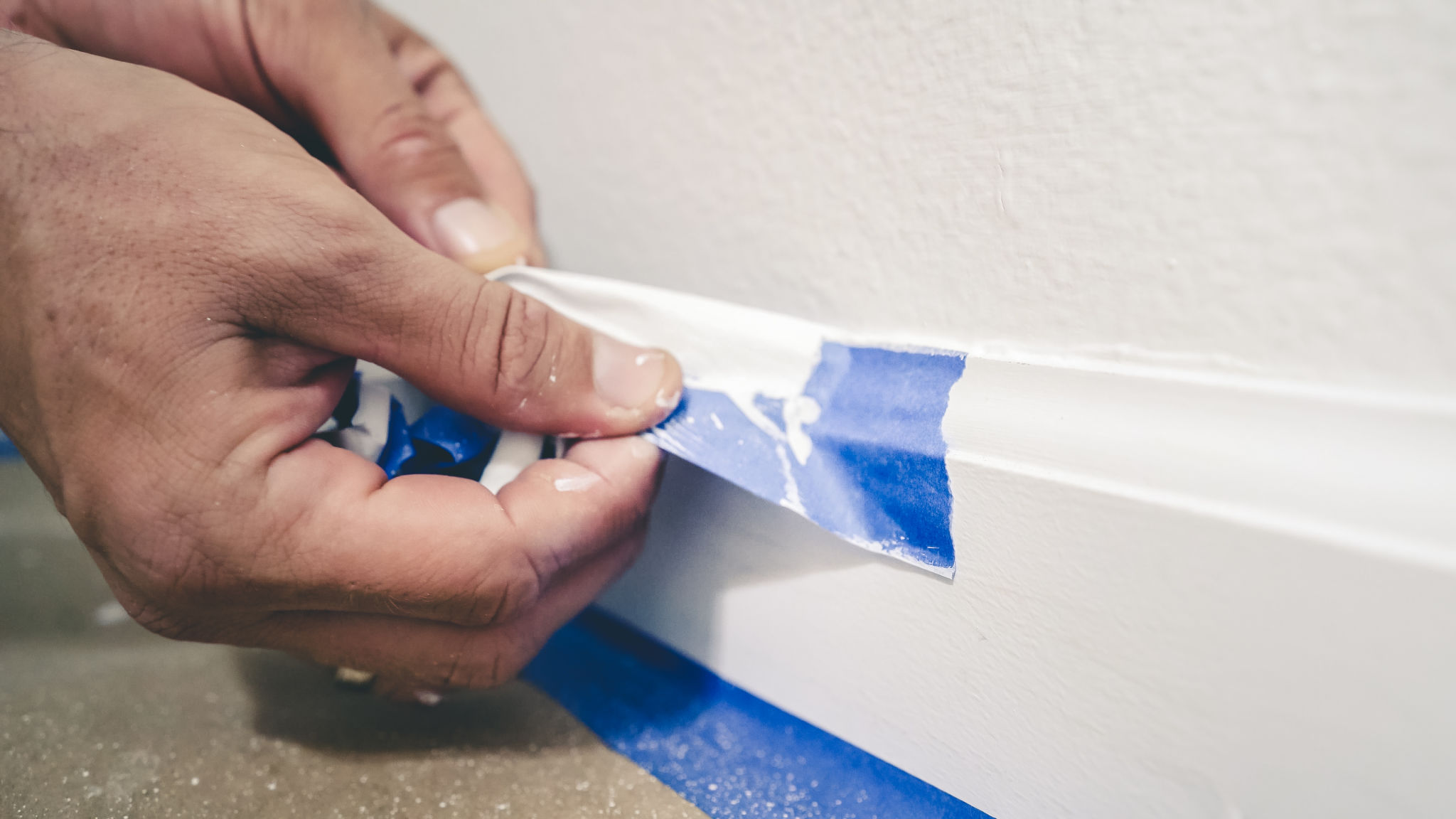 Removing masking tape from moulding. A painter pulls of blue painter's tape from the wall to reveal a clean edge baseboard. Removing masking tape from moulding. A painter pulls of blue painter's tape from the wall to reveal a clean edge baseboard.