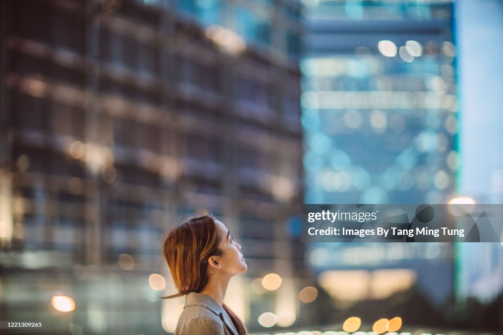 Portrait of Asian woman looking up to sky with confidence against illuminated city buildings at dawn