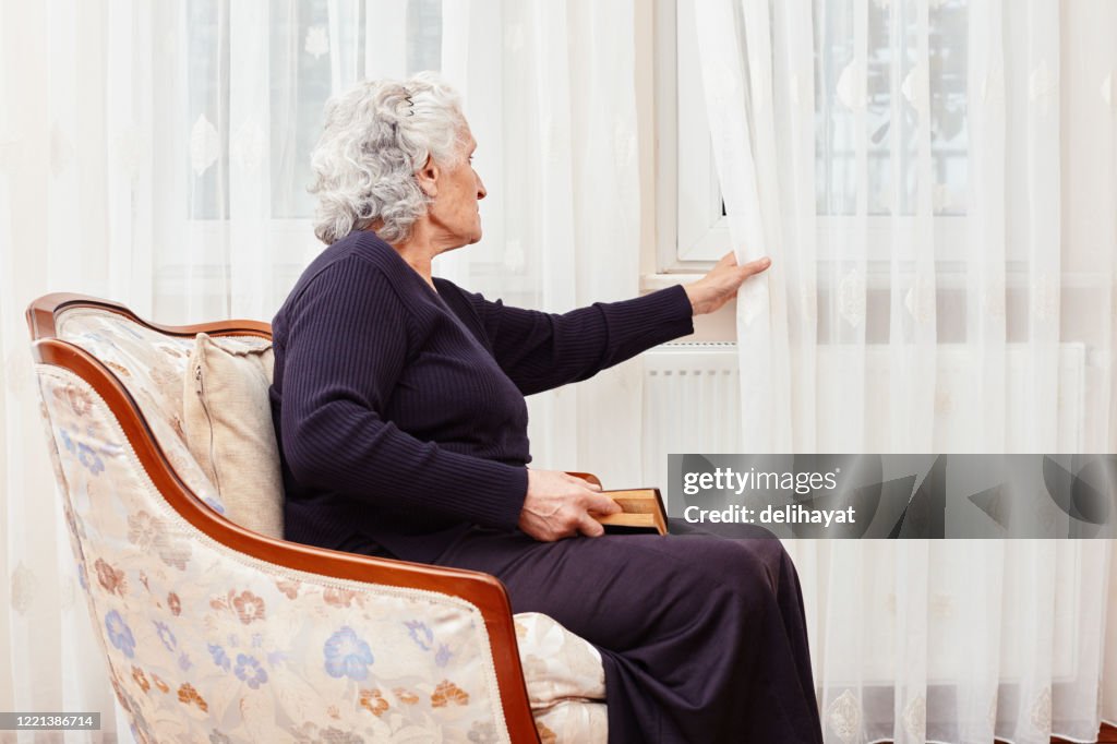 Mujer anciana sentada sola con un libro en la mano y mira tristemente fuera de la ventana