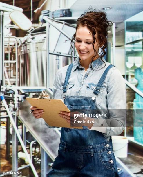 young women entrepreneur at the warehouse wearing bib overalls and holding clipboard - contract manufacturing stock pictures, royalty-free photos & images