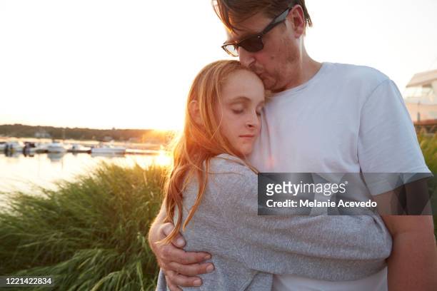 father and daughtersunset hitting the back of their head standing infront of grass on beach water dock in background looking away from camera father is leaning and hugging girl daughters arms are wraped around her