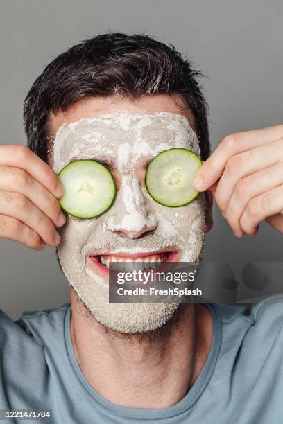 men's face care: smiling man with a moisturizing mask putting slices of cucumber over his eyes to ease puffiness - men facial stock pictures, royalty-free photos & images