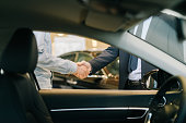 Buyer of car shaking hands with seller in auto dealership, view from interior of car.