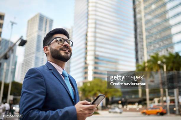 portrait of optimistic hispanic businessman with smart phone - looking up at buildings stock pictures, royalty-free photos & images
