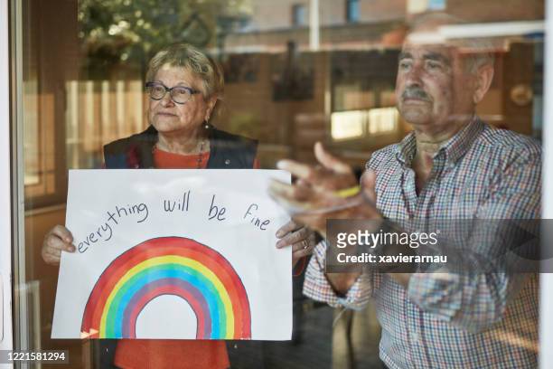 senior couple applauding and showing a hand drawn rainbow at home in quarantine - state of emergency sign stock pictures, royalty-free photos & images