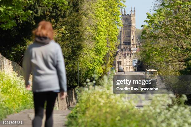 mujer caminando hacia el centro de la ciudad de cirencester iglesia parroquial en cotswolds, gloucestershire, asomando entre los árboles y el follaje y un brillante día de primavera - cirencester fotografías e imágenes de stock