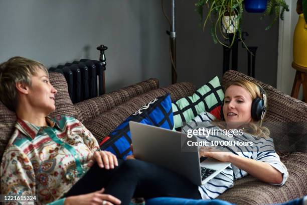 lesbian couple relaxing on sofa one using laptop - downloading stock pictures, royalty-free photos & images