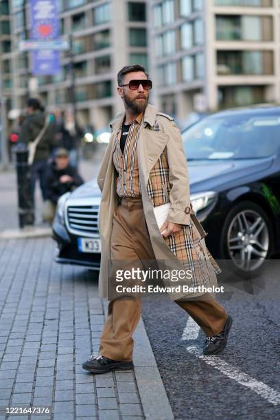 Guest wears sunglasses, a Burberry checked trench beige coat, brown pants, striped shirt, shoes, during London Fashion Week February 2020 on February...