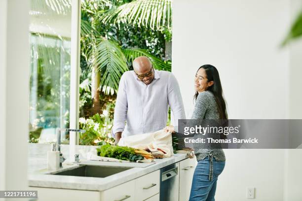 laughing couple putting away groceries in kitchen - friendship home ownership stock pictures, royalty-free photos & images