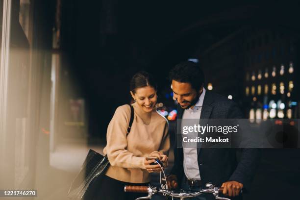 smiling woman showing smart phone to male friend with bicycle while standing in city at night - fahrrad nacht stock-fotos und bilder
